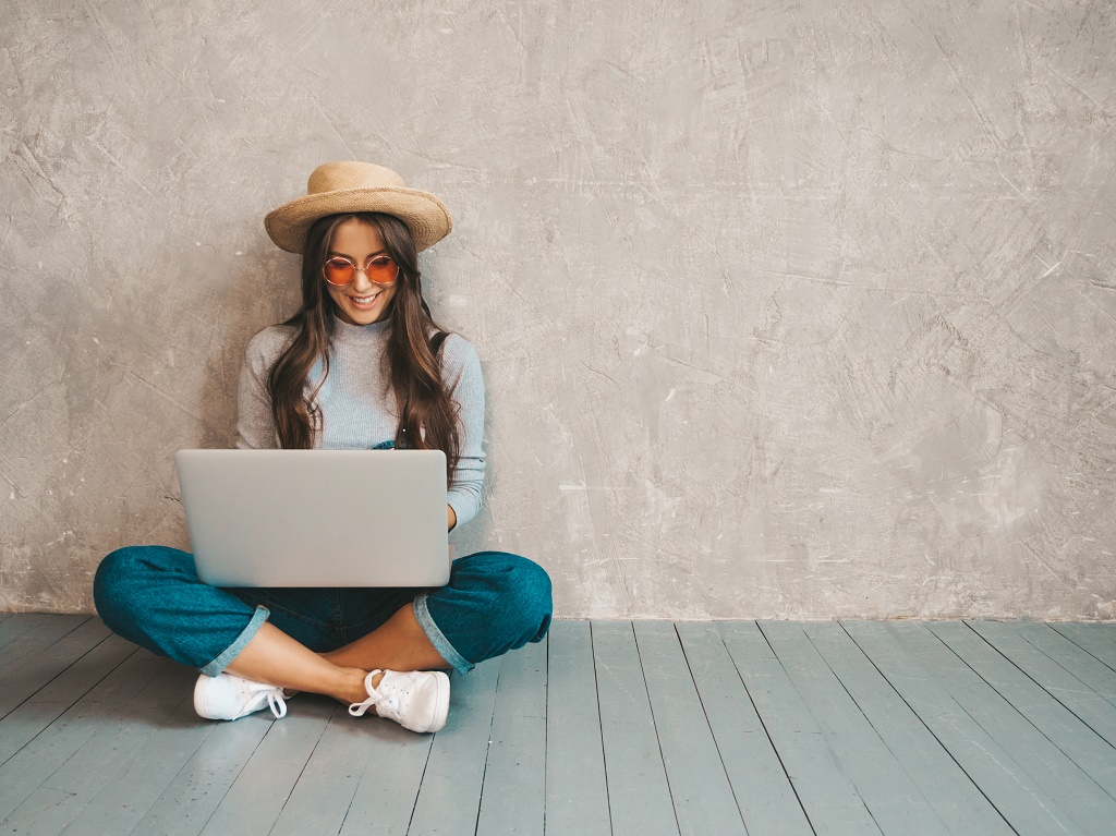 Portrait of creative young smiling woman in sunglasses. Beautiful girl sitting on the floor near gray wall. Model using notebook. Female dressed in hipster clothes and hat