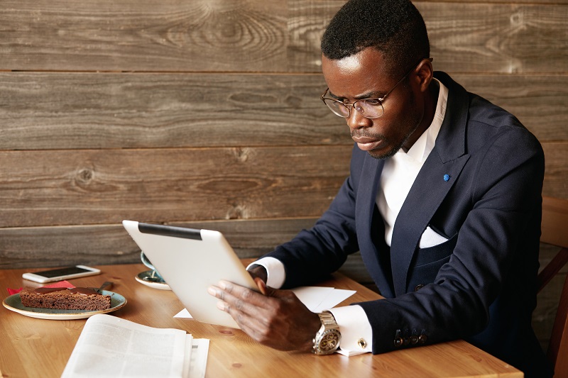 Serious young African American businessman holding digital tablet, looking at the screen with concentrated expression while filling in some papers, sitting at a cafe, having coffee during lunch break