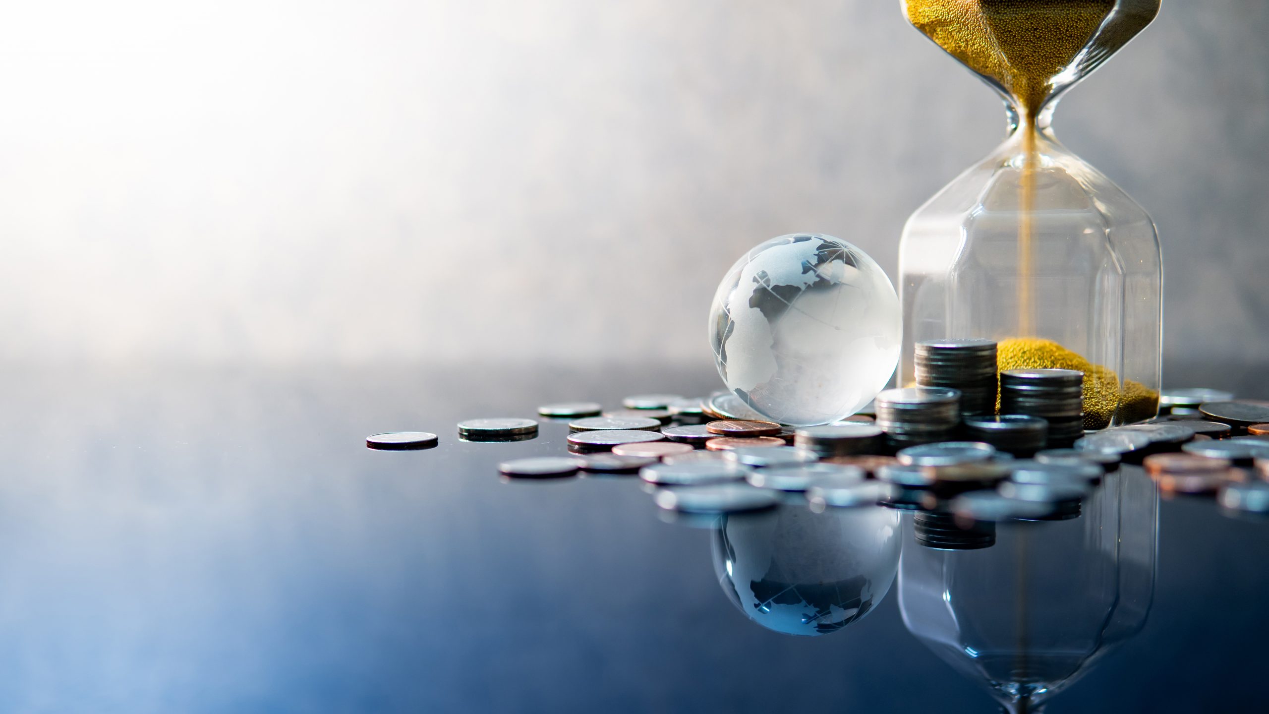 Global business investment growth. Wealth concept. Gold sand running through the shape of modern hourglass with world globe crystal glass and US dollar coins on blue reflective table.
