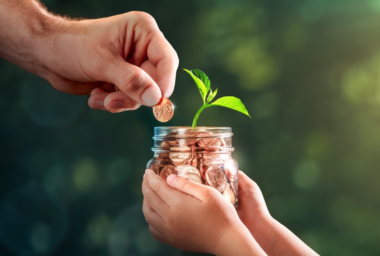 Hand Of Father Adding Penny To Child's Coin Jar With Plant Growing Out Of It - College Fund / Investing In Our Children's Future Concept