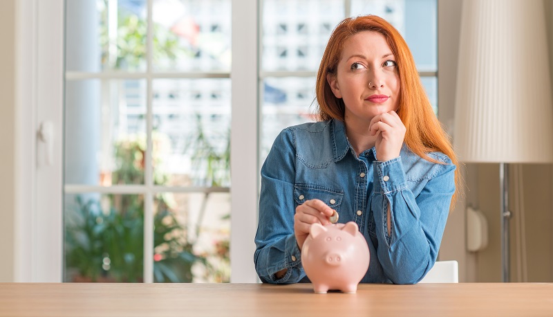 Redhead woman saves money in piggy bank at home serious face thinking about question, very confused idea