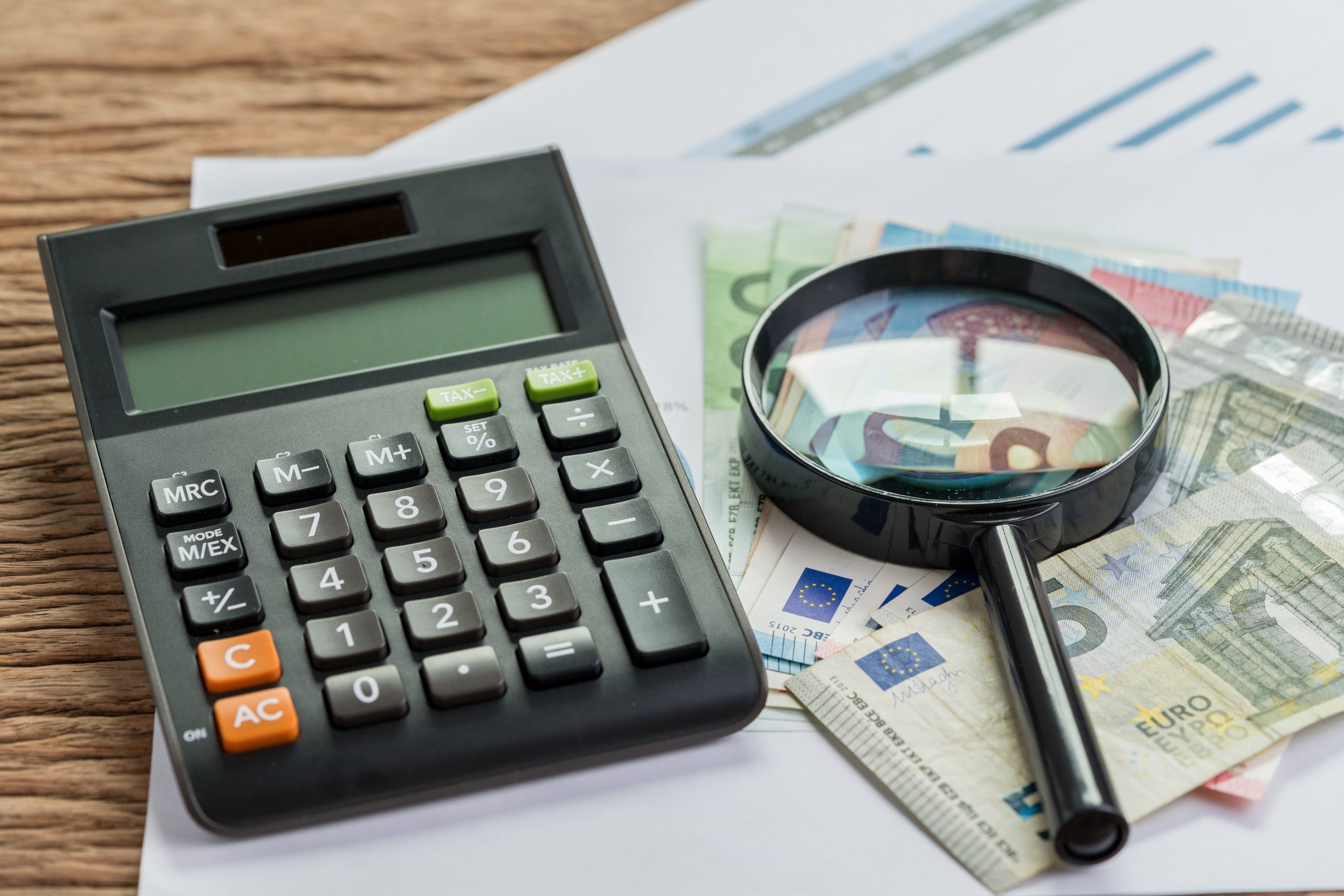 Tax or searching for yield concept, calculator with magnifier glass on pile of Euro banknotes on graph and chart performance print paper on wood table