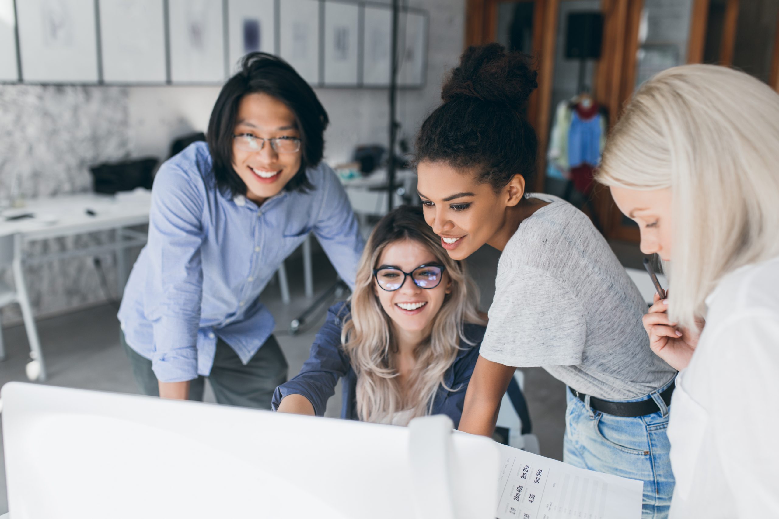 Long-haired blonde secretary in glasses laughing while looking at computer with asian programmer and african girl. Indoor portrait of chinese office worker and his colleagues with laptop foreground.
